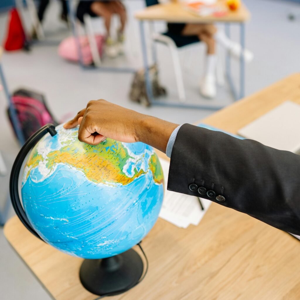 pexels-photo-8617981-8617981 A teacher's hand points at a globe in a classroom while students are present.